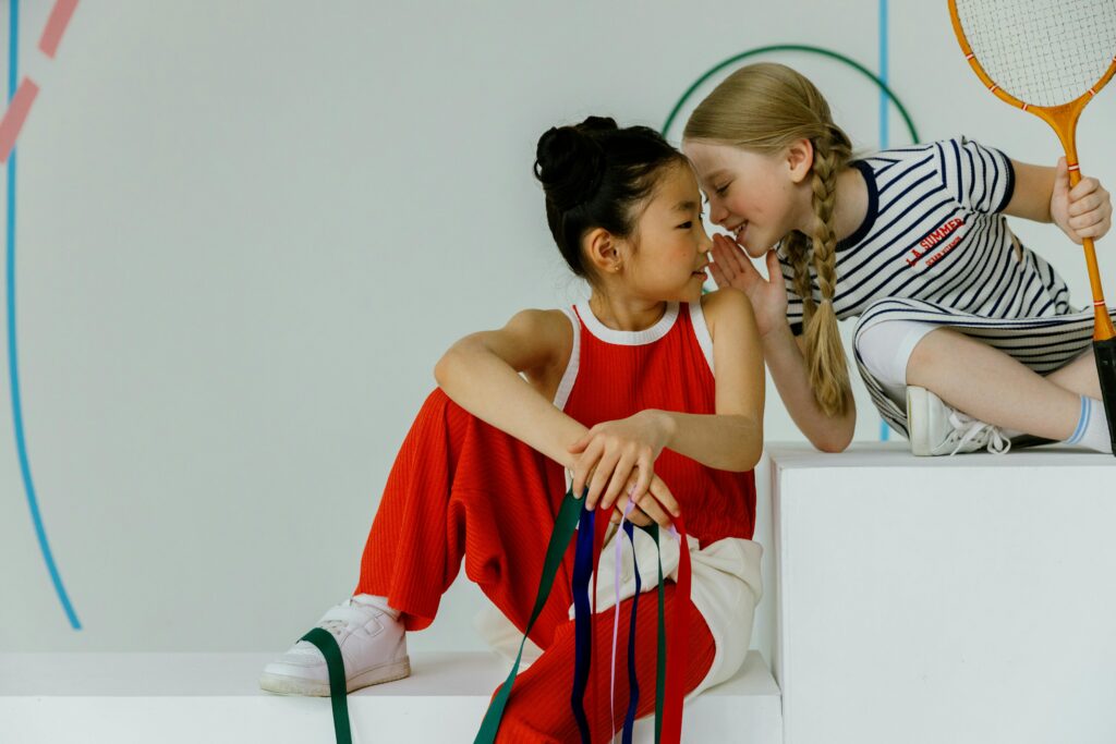 Two young girls whispering together, one holding a tennis racket, dressed in sportswear indoors.